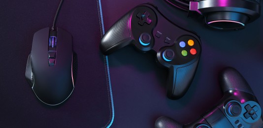 Photo of Computer mouse, mousepad and game controllers in neon lights on black background, top view