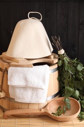 Sauna equipment. Bucket, ladle, felt wool hat, birch whisk and towel on bamboo bench indoors