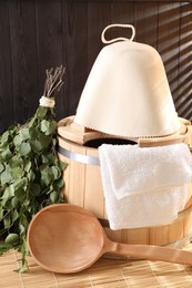 Photo of Sauna equipment. Bucket, ladle, felt wool hat, birch whisk and towel on bamboo bench indoors