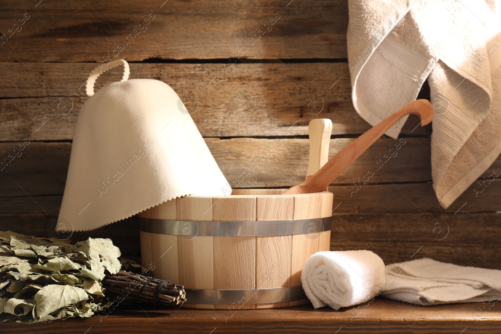 Photo of Sauna equipment. Bucket with ladle, felt wool hat, oak whisk and towels on wooden table indoors