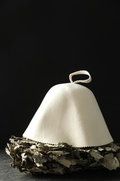 Photo of Sauna equipment. Oak whisk and felt wool hat on dark textured surface against black background, space for text