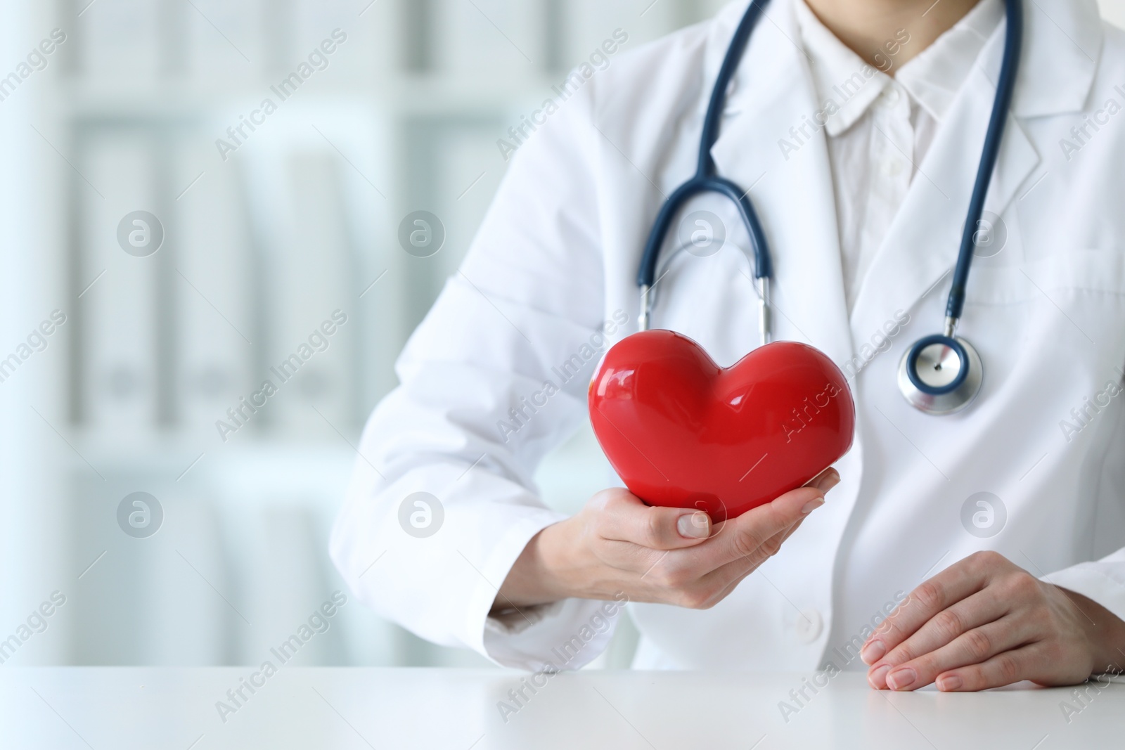 Photo of Doctor with red heart at table in clinic, closeup. Space for text