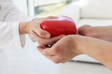 Doctor giving red heart to patient at white table in clinic, closeup