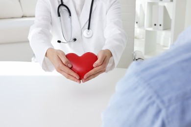 Doctor showing red heart to patient at white table in clinic, closeup