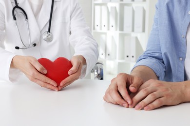 Doctor showing red heart to patient at white table in clinic, closeup