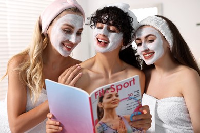 Photo of Happy friends with facial masks reading magazine in bathroom. Spa party