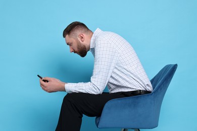 Photo of Man with poor posture sitting on chair and using smartphone against light blue background