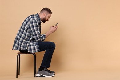 Photo of Man with poor posture sitting on chair and using smartphone against pale orange background, space for text