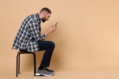 Man with poor posture sitting on chair and using smartphone against pale orange background, space for text