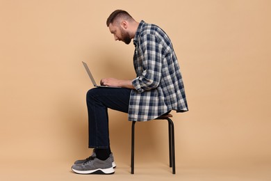 Man with poor posture sitting on chair and using laptop against pale orange background