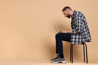 Photo of Man with poor posture sitting on chair and using laptop against pale orange background, space for text