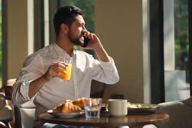 Happy man having tasty breakfast while talking on smartphone in cafe, space for text