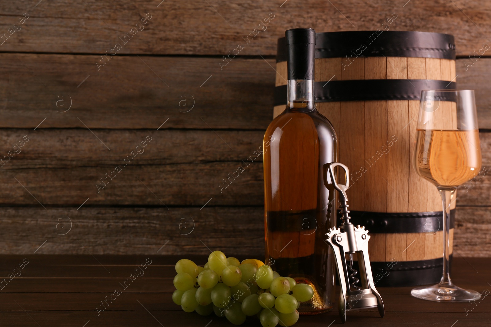 Photo of Glass of wine, bottle, wing corkscrew, barrel and grapes on wooden table, space for text