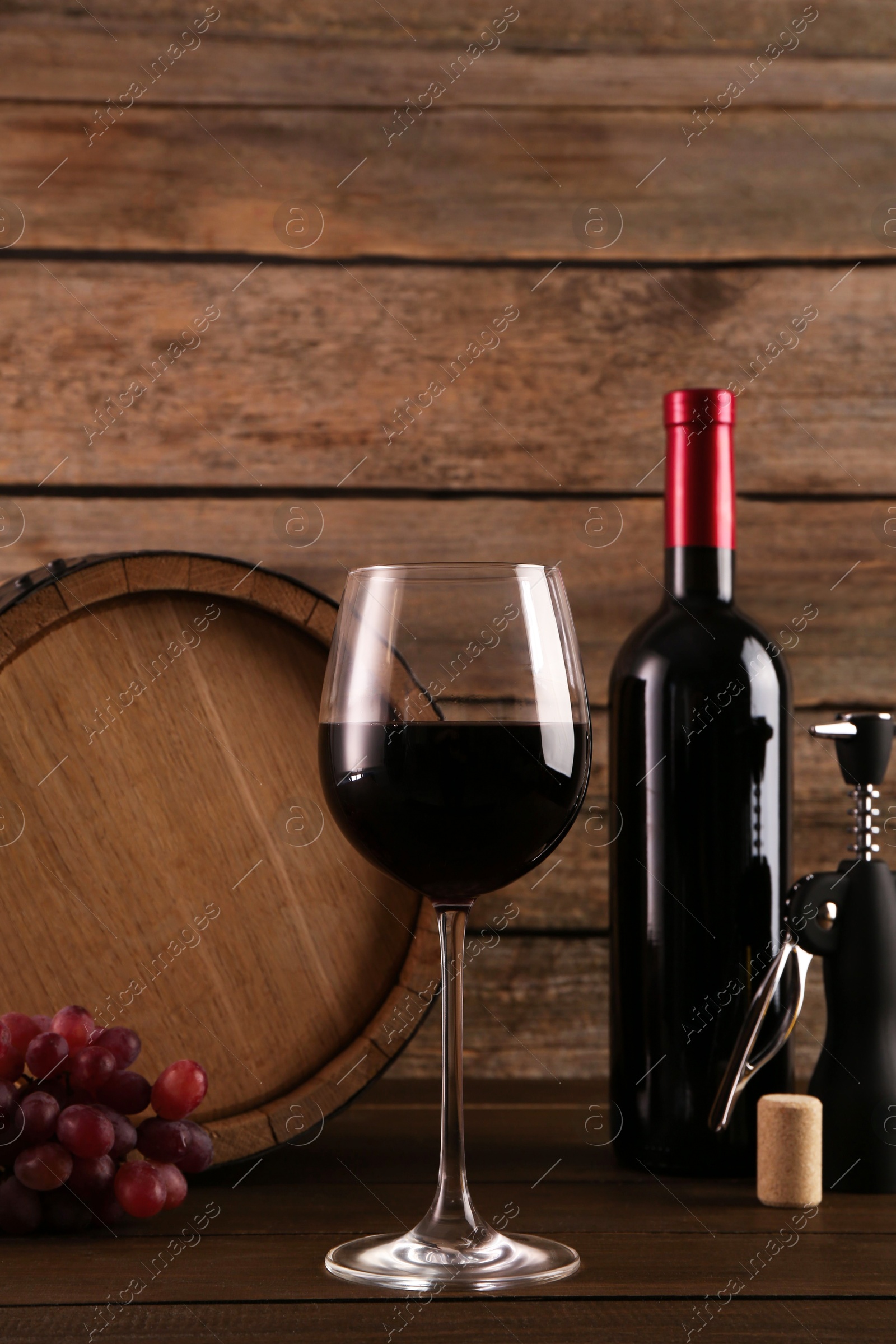 Photo of Glass of wine, bottle, wing corkscrew, barrel and grapes on wooden table