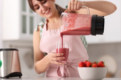 Photo of Woman pouring fresh smoothie from blender cup into glass in kitchen, selective focus