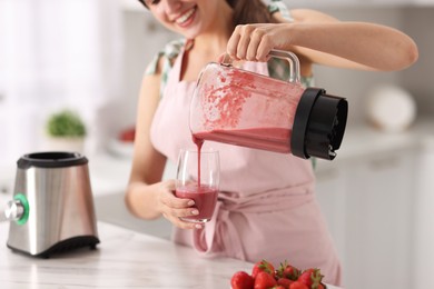Photo of Woman pouring fresh smoothie from blender cup into glass at white marble table in kitchen, closeup