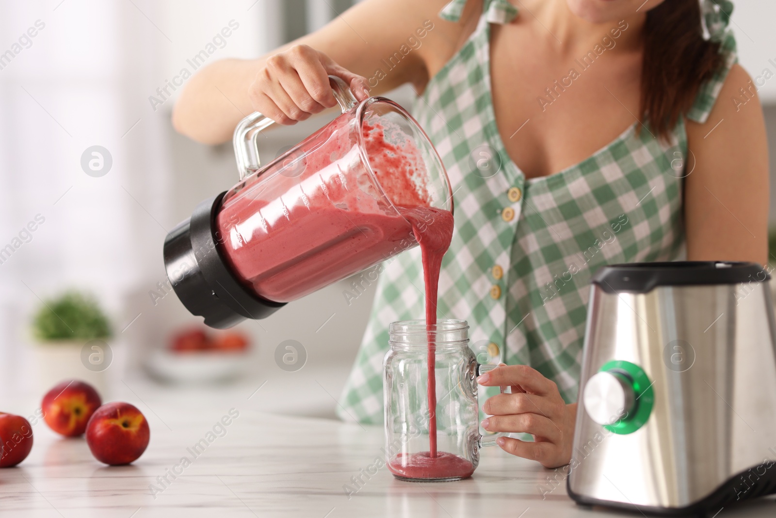 Photo of Woman pouring fresh smoothie from blender cup into glass at white marble table in kitchen, closeup