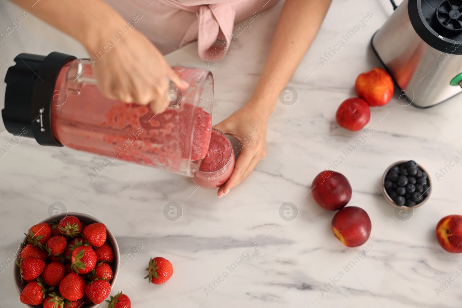 Photo of Woman pouring fresh smoothie from blender cup into glass at white marble table in kitchen, top view