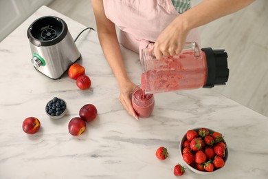 Photo of Woman pouring fresh smoothie from blender cup into glass at white marble table in kitchen, closeup