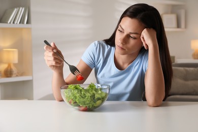 Eating disorder. Sad woman holding fork with tomato over bowl at table indoors