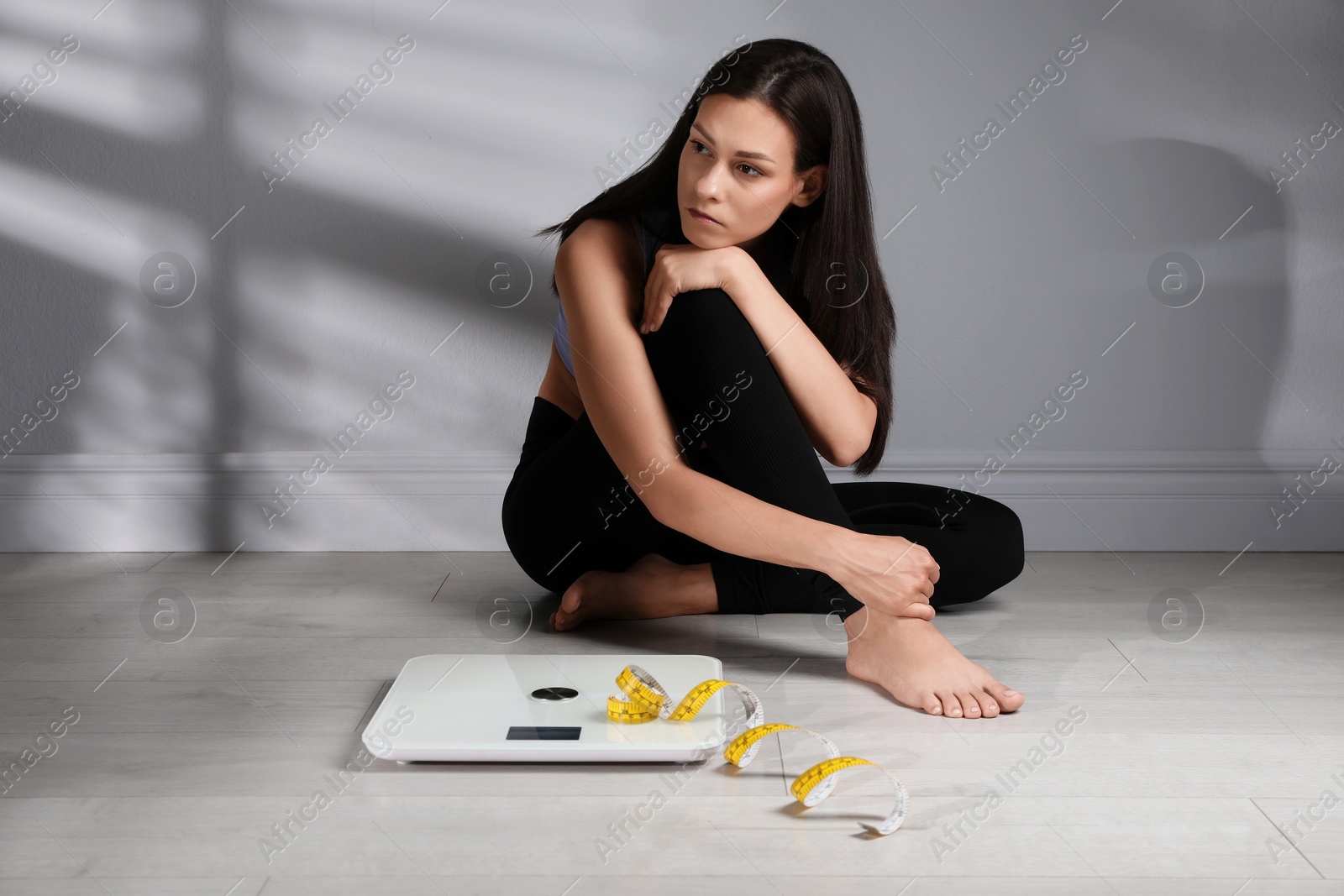 Photo of Eating disorder. Sad woman sitting near scale and measuring tape on floor indoors