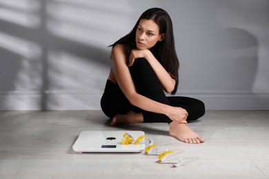 Eating disorder. Sad woman sitting near scale and measuring tape on floor indoors