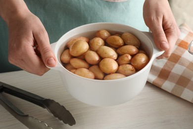 Photo of Woman holding pot with raw potatoes at white wooden table, closeup