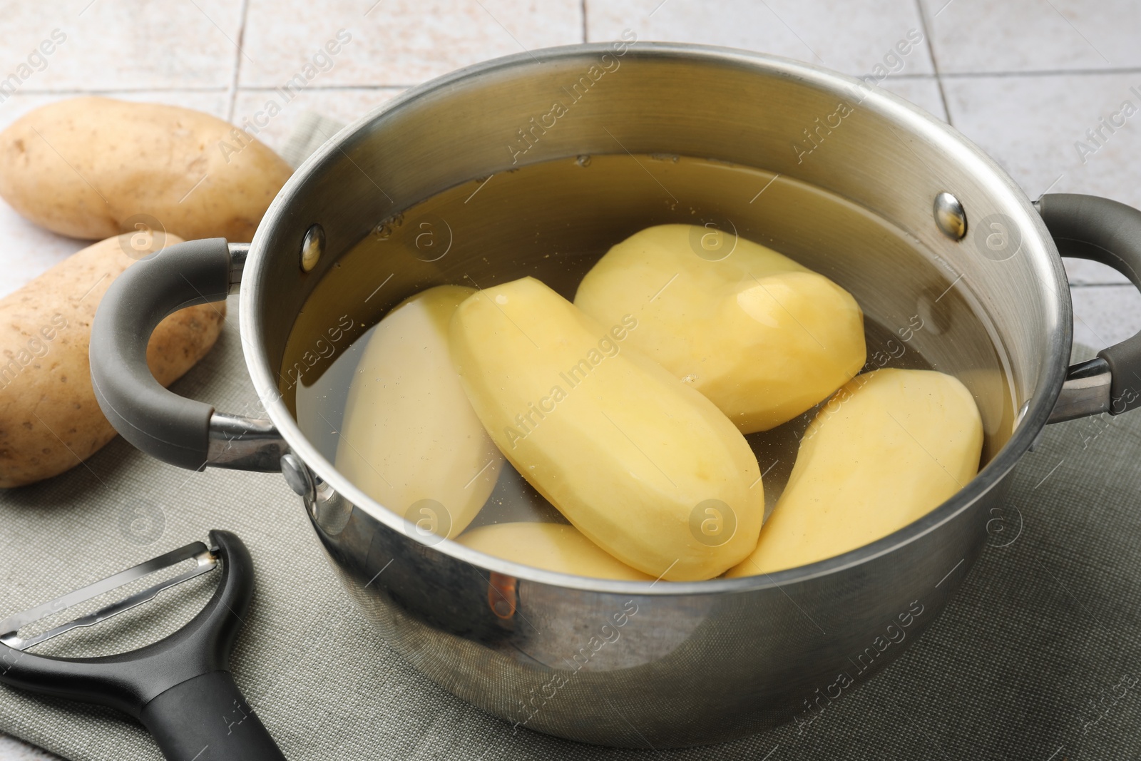 Photo of Raw potatoes in pot with water and peeler on table