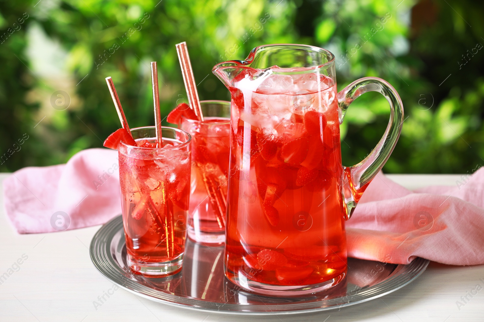 Photo of Tasty strawberry lemonade in jug and glasses on white table against blurred green background