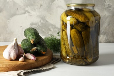 Pickles in jar, fresh cucumbers, garlic and dill on white table