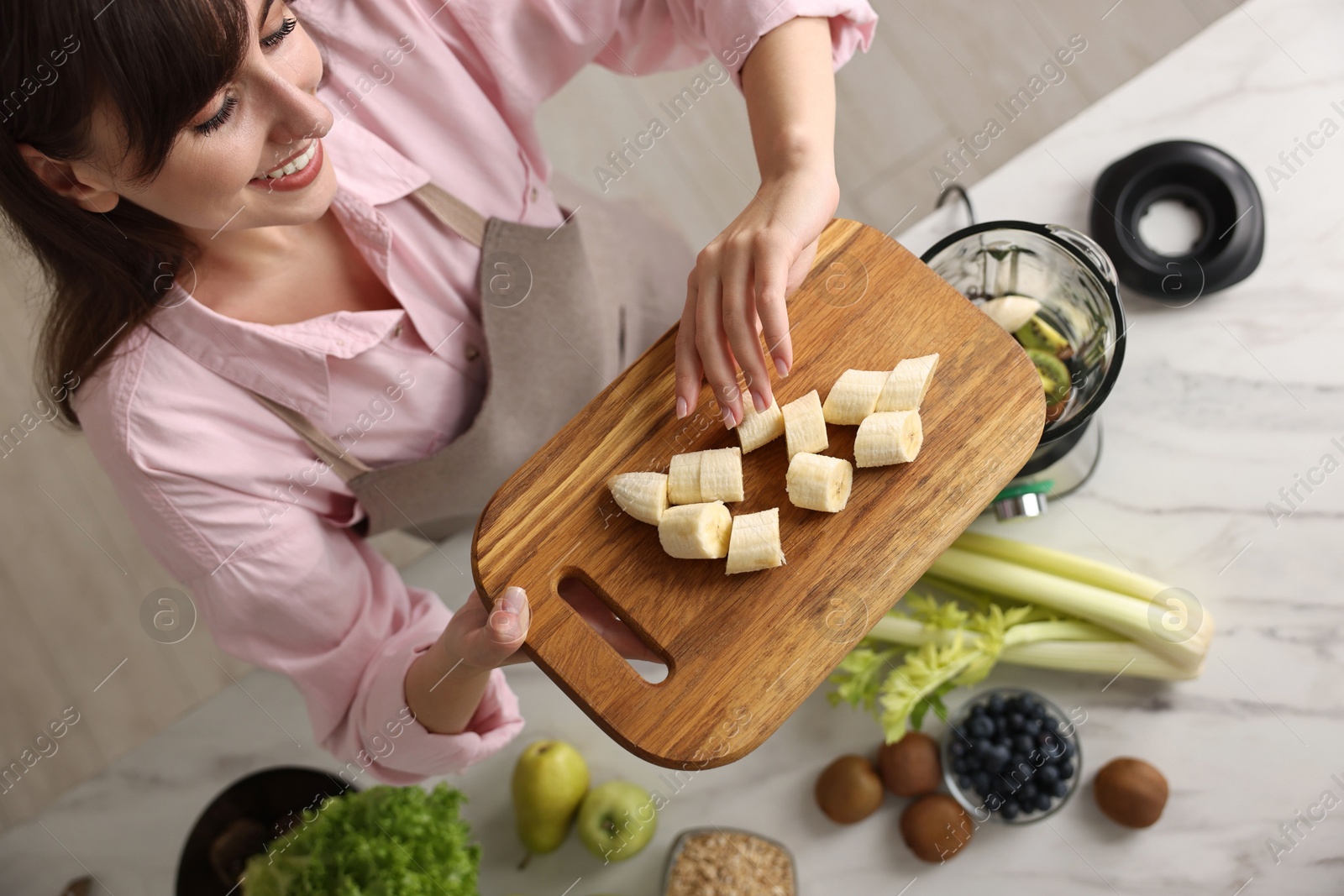 Photo of Young woman making delicious smoothie with blender at white marble table in kitchen, top view