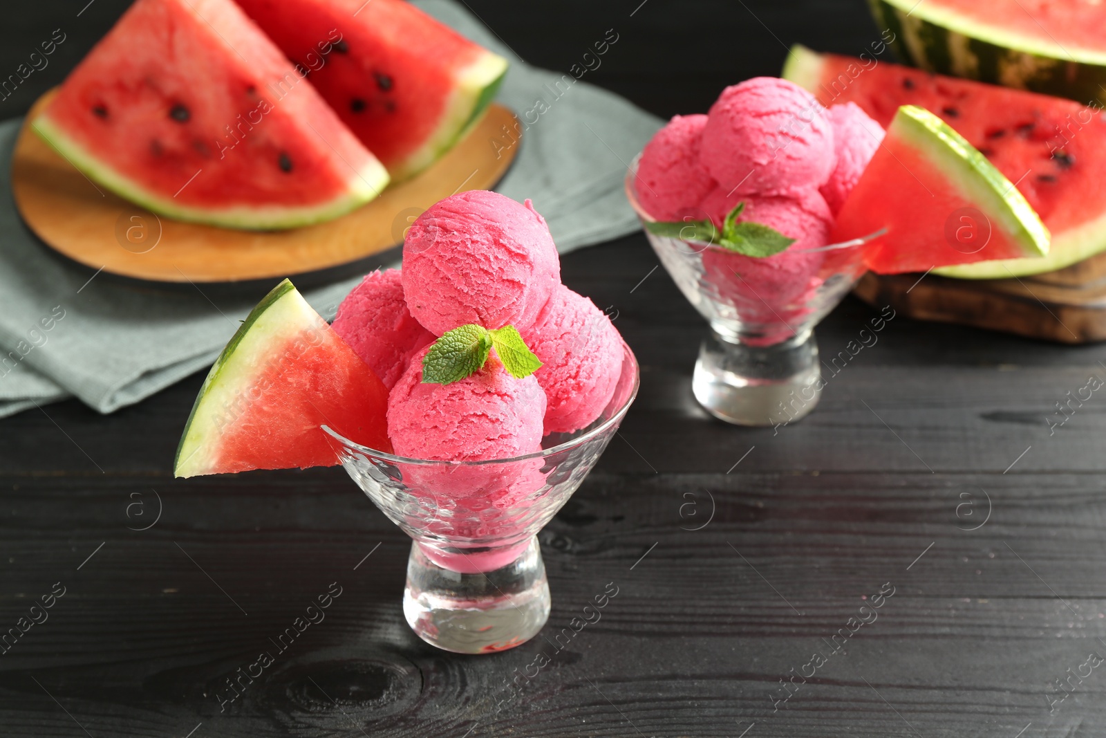 Photo of Scoops of tasty watermelon sorbet with mint in glass dessert bowls and fresh fruit on black wooden table