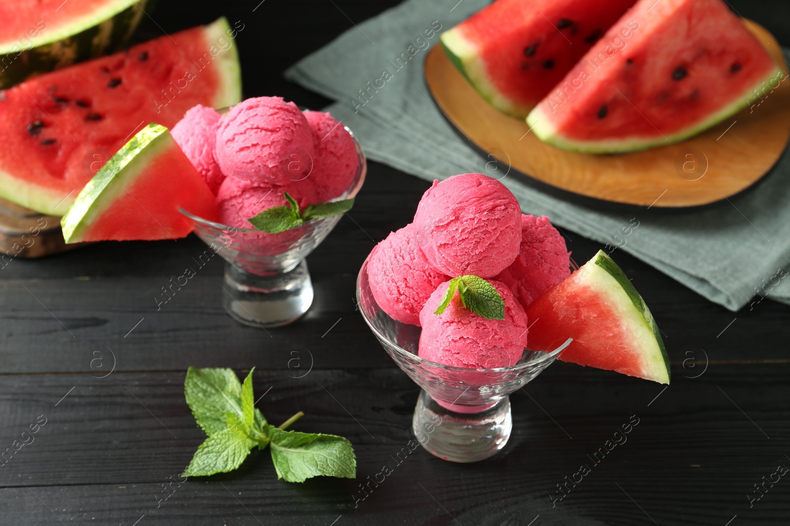 Photo of Scoops of tasty watermelon sorbet with mint in glass dessert bowls and fresh fruit on black wooden table