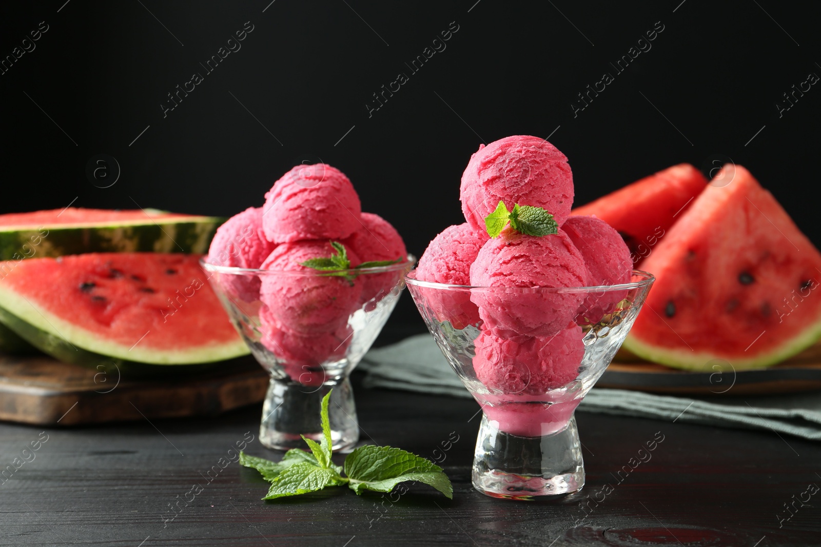 Photo of Scoops of tasty watermelon sorbet with mint in glass dessert bowls on black wooden table