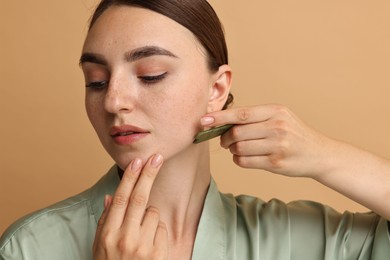 Photo of Beautiful young woman doing facial massage with gua sha tool on beige background