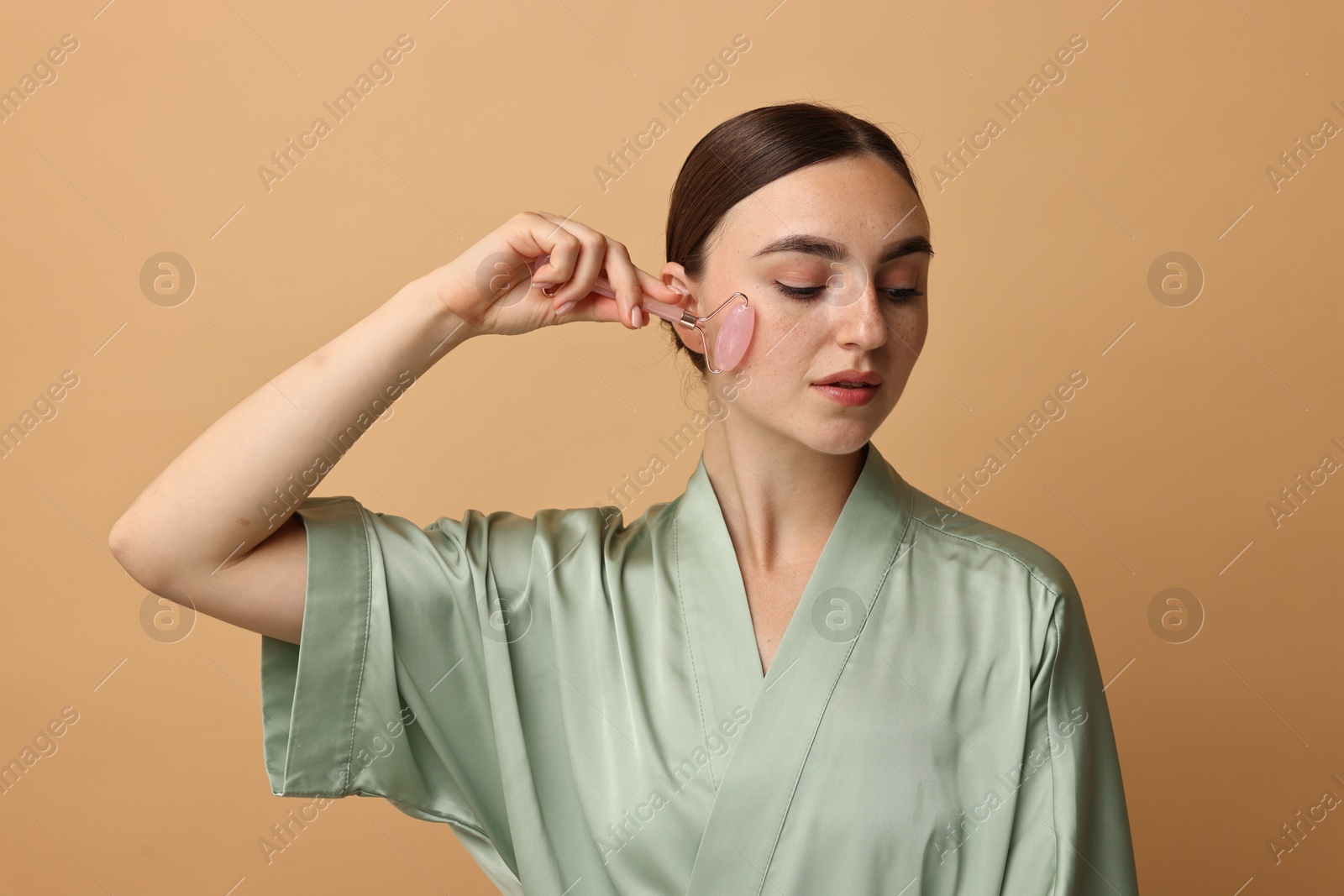 Photo of Beautiful young woman doing facial massage with roller on beige background