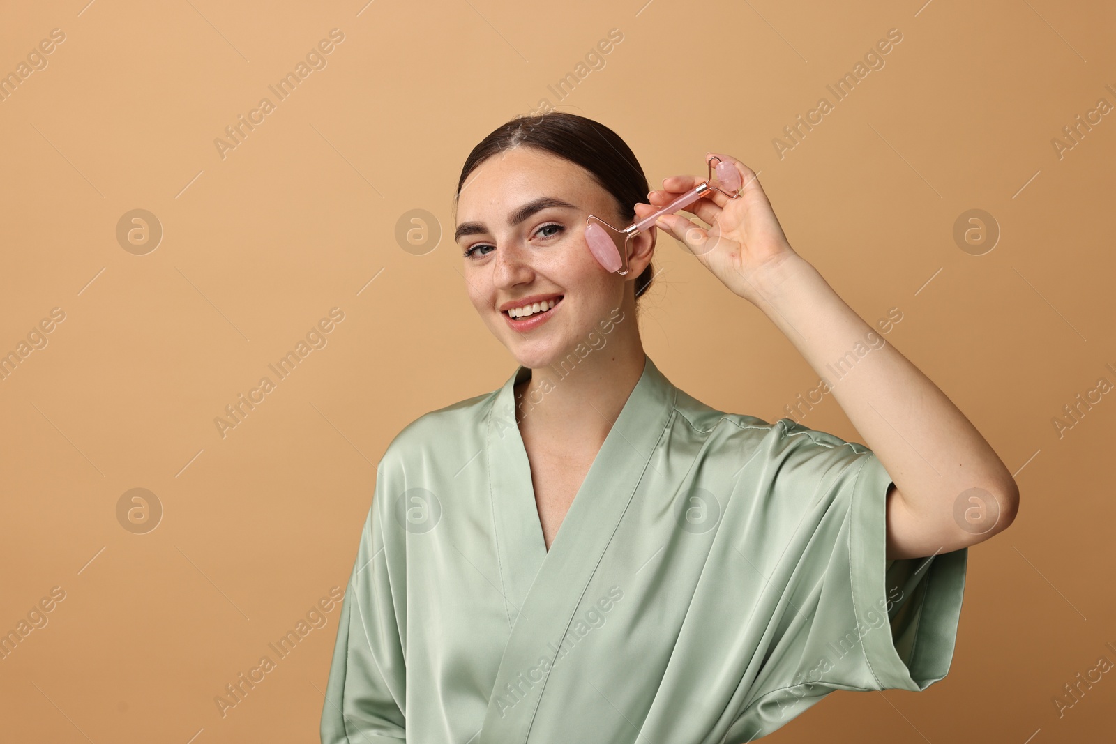 Photo of Beautiful young woman doing facial massage with roller on beige background