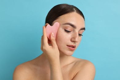 Photo of Beautiful young woman doing facial massage with gua sha tool on light blue background