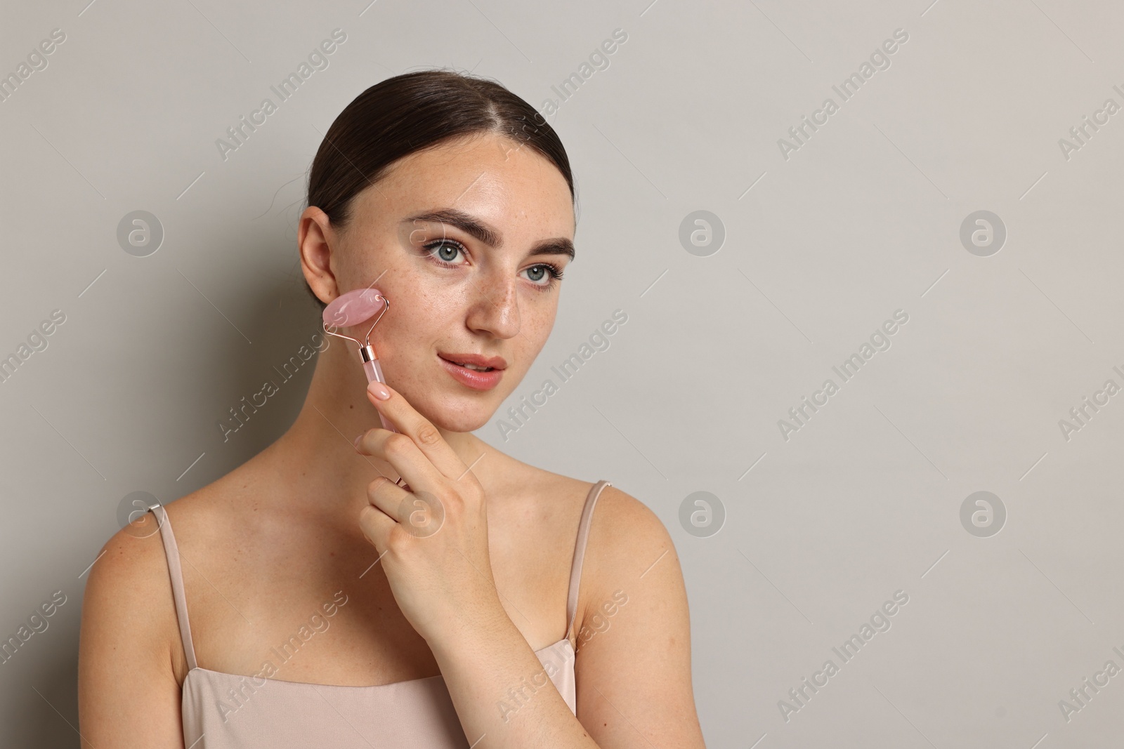 Photo of Beautiful young woman doing facial massage with roller on grey background, space for text