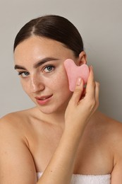 Beautiful young woman doing facial massage with gua sha tool on grey background