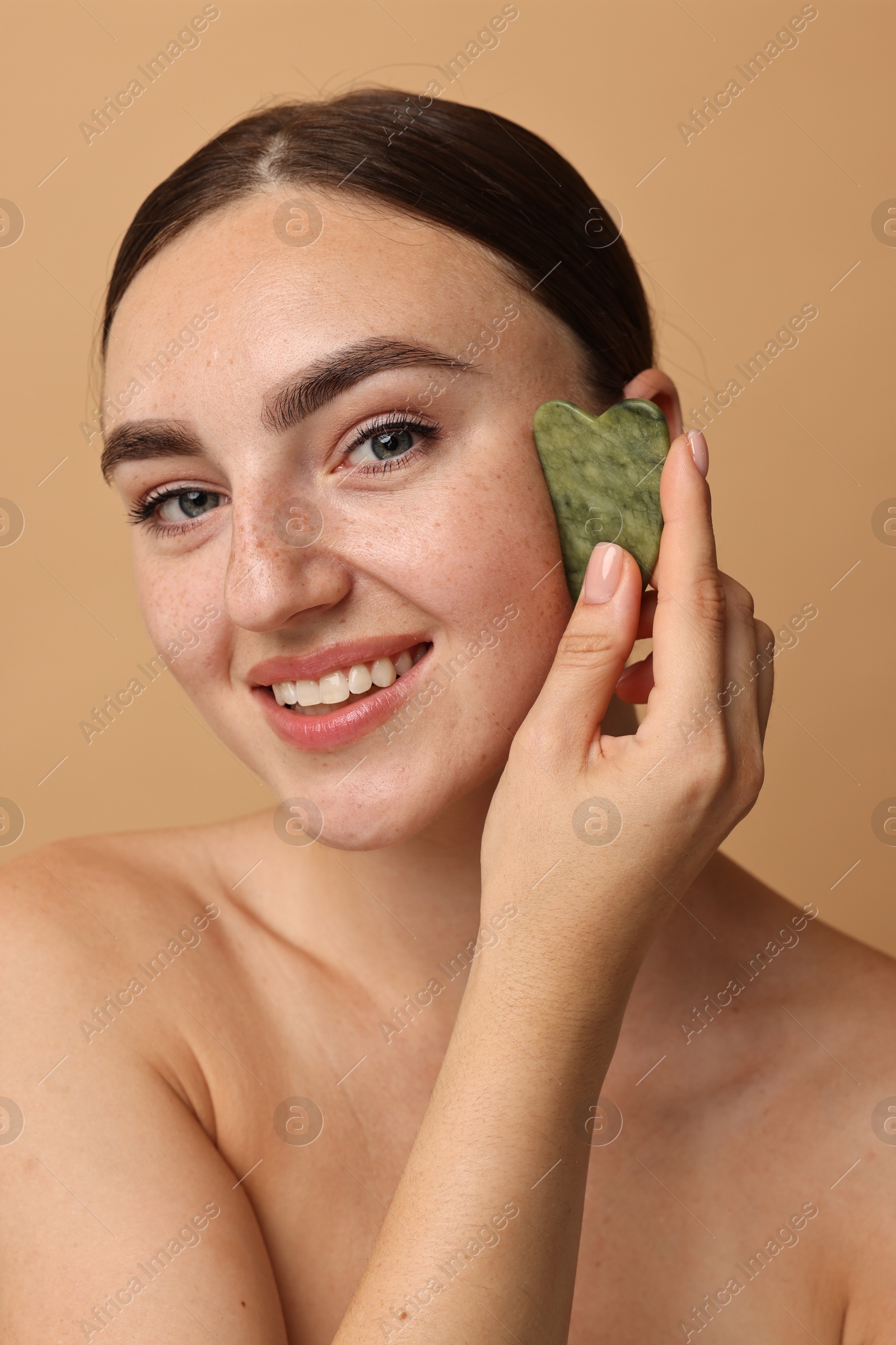 Photo of Beautiful young woman doing facial massage with gua sha tool on beige background