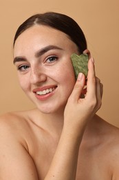 Beautiful young woman doing facial massage with gua sha tool on beige background