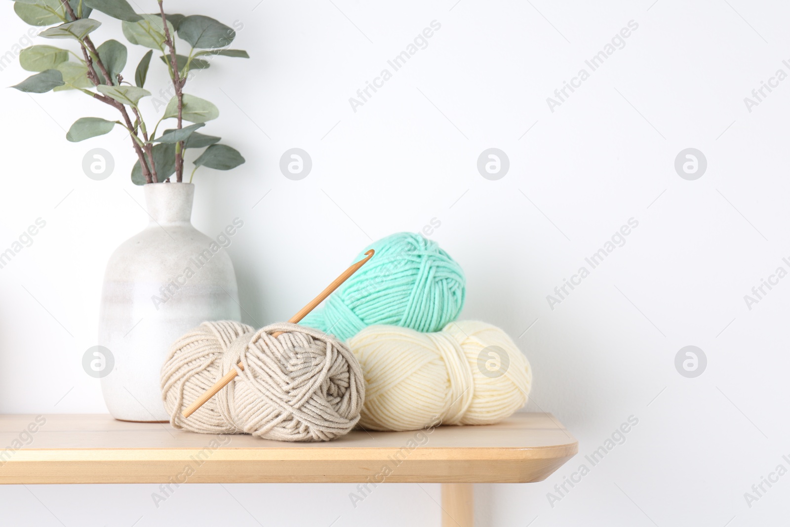 Photo of Skeins of yarn, eucalyptus in vase and crochet hook on wooden shelf. Space for text