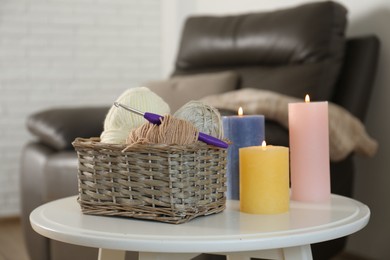 Photo of Wicker basket with different yarns and crochet hook near burning candles on table indoors