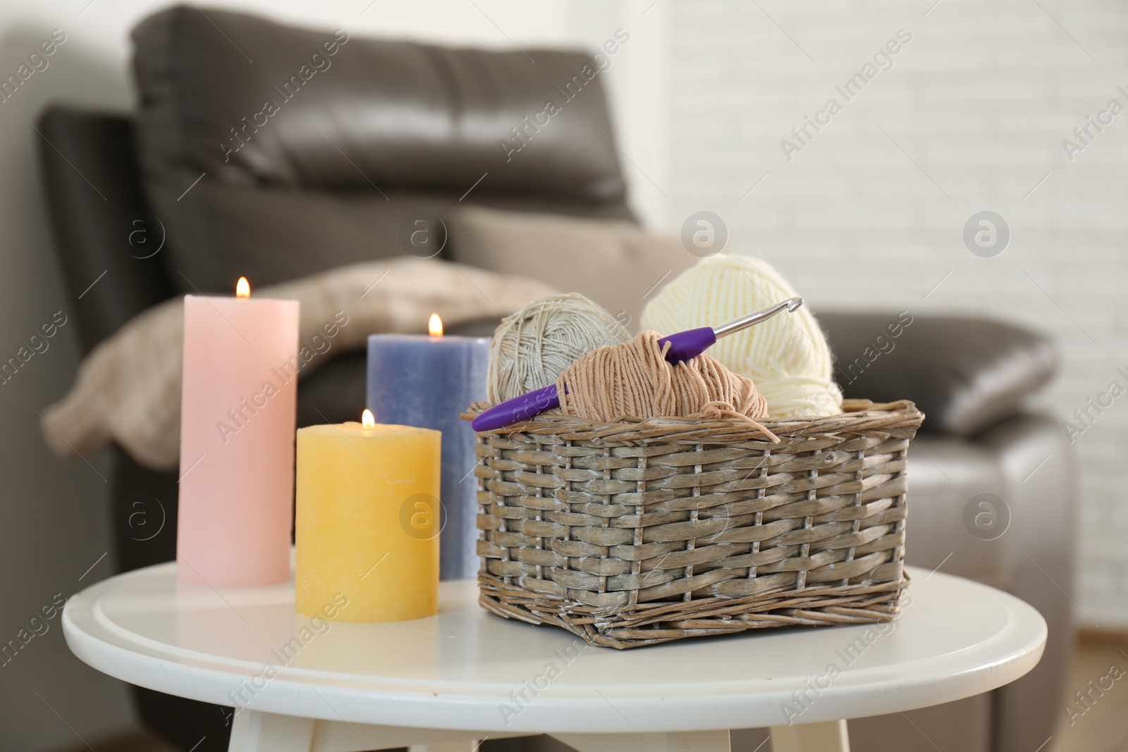 Photo of Wicker basket with different yarns and crochet hook near burning candles on table indoors