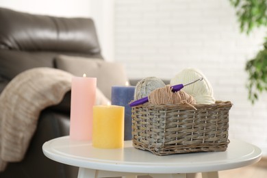 Photo of Wicker basket with different yarns and crochet hook near burning candles on table indoors