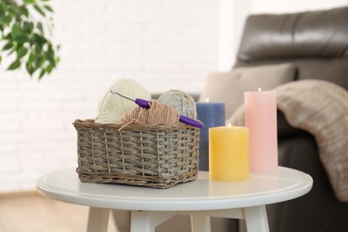 Photo of Wicker basket with different yarns and crochet hook near burning candles on table indoors