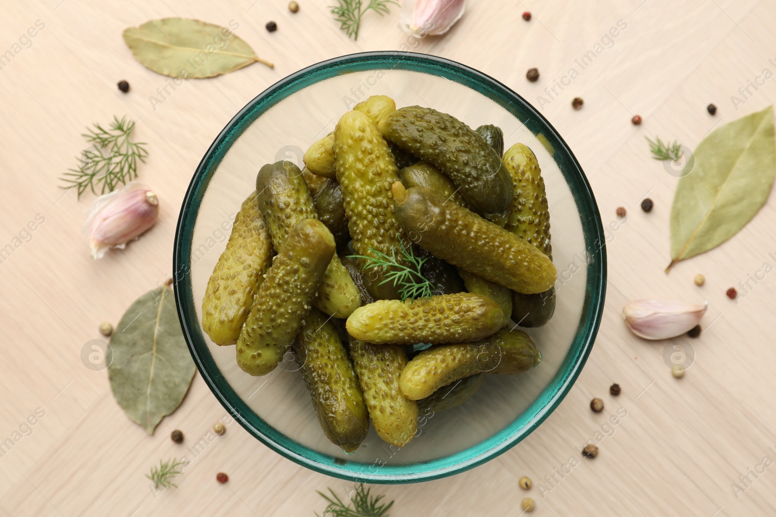 Photo of Pickled cucumbers in bowl surrounded by spices on light wooden table, flat lay