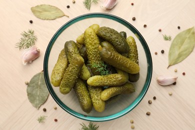 Pickled cucumbers in bowl surrounded by spices on light wooden table, flat lay