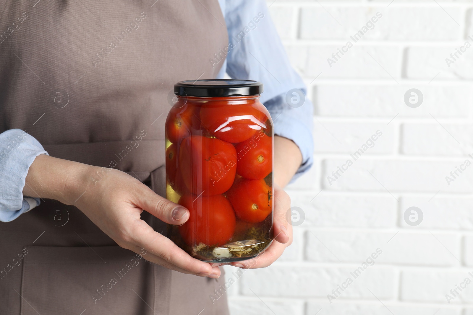 Photo of Woman holding jar with tasty pickled tomatoes against white brick wall, closeup. Space for text